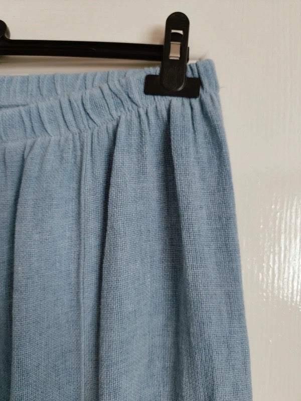 A close-up of a light blue textured fabric garment, likely a skirt or drape, hanging from a black hanger with a black clip against a plain off-white wall.